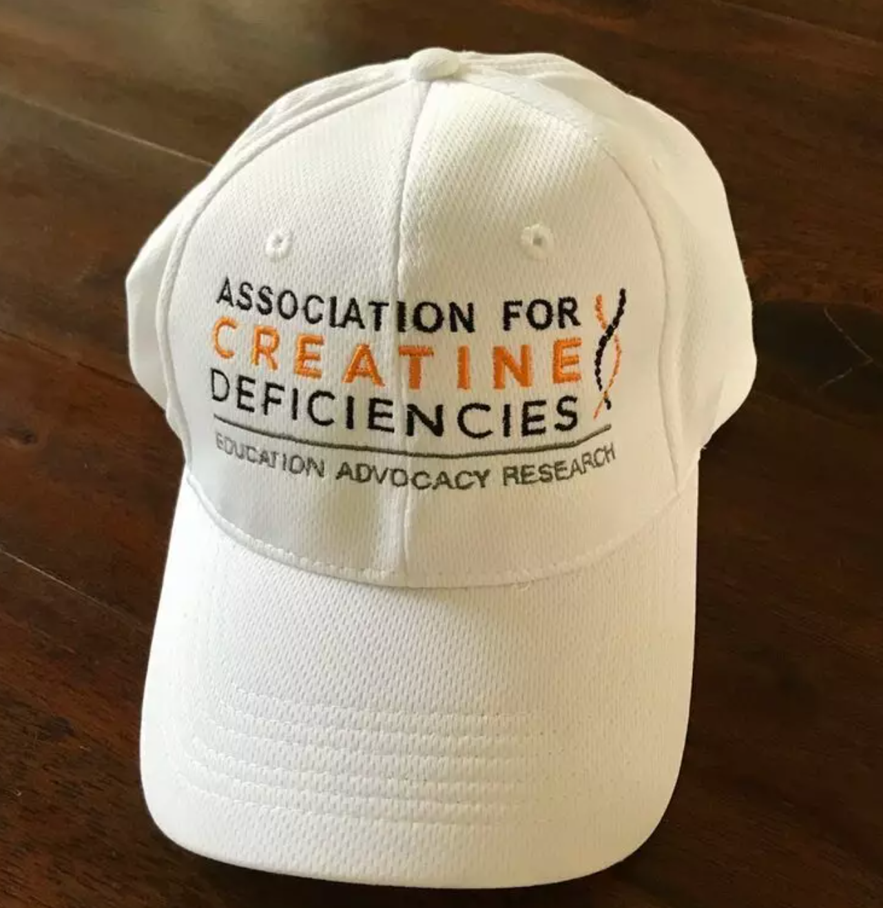 White hat on a wood background. Hat says Association for Creatine Deficiencies on it.